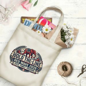 Natural canvas tote bag with a "Hold The Line Red Wave 2026" graphic print, shown open with magazines and flowers inside, placed on a white wooden background.