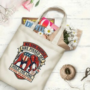Natural canvas tote bag with a "We The People Are Pissed Off Red Wave 2026" graphic print, shown open with magazines and flowers inside, placed on a white wooden background.