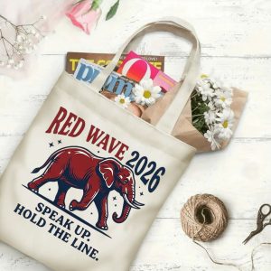 Natural canvas tote bag with "Red Wave 2026" graphic print, shown open with magazines and flowers inside, placed on a white wooden background.