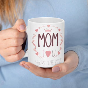 A white 11oz ceramic coffee mug held in hands, featuring "MOM" in bold mauve letters, a pink heart wreath with a crown, "I ❤️ U", and "You're the best" below.