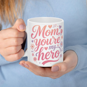 A white 11oz ceramic coffee mug held in hands, featuring elegant pink-to-orange cursive text reading "Mom, you're my hero" surrounded by delicate flowers and hearts.