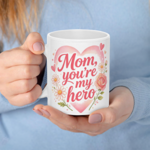 A white 11oz ceramic coffee mug held in hands, featuring "Mom, you're my hero" in coral cursive text inside a pink watercolor heart, surrounded by daisies, roses, and hearts.