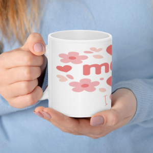 A white ceramic coffee mug held in hands, featuring a pink floral and heart wreath design with "mom" and "I ❤️ U".