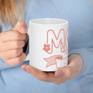 A white 11oz ceramic coffee mug held in hands, featuring a cute coral doodle design where the "O" in "MOM" is replaced by a pink heart, with flowers and a banner reading "I LOVE YOU".