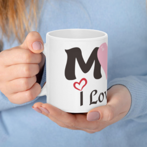 A white 11oz ceramic coffee mug held in hands, featuring "MOM" in bold black letters with a pink heart in place of the "O", plus "I Love You" written below with a small red heart accent.