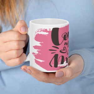 A white 11oz ceramic coffee mug held in hands, featuring "Love my mom" in bold black cursive text over a pink brushstroke background with heart accents.