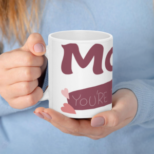 A white 11oz ceramic coffee mug held in hands, featuring "MOM" in large mauve letters with a banner reading "You're the best" and small pink heart accents.