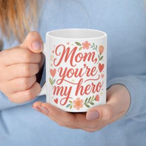 A white 11oz ceramic coffee mug held in hands, featuring elegant coral cursive text reading "Mom, you're my hero" surrounded by colorful flowers and hearts.