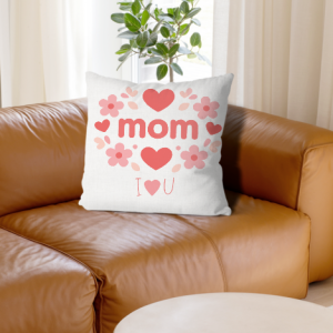 A square white throw pillow with a pink floral and heart wreath design, reading "mom" and "I ❤️ U", placed on a brown leather sofa.