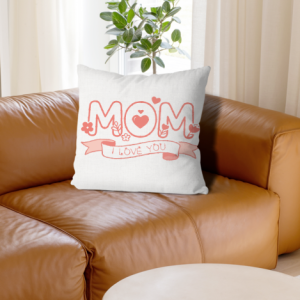 A square white throw pillow with a coral-pink "MOM" doodle design, hearts, flowers, and a banner reading "I LOVE YOU", placed on a brown leather sofa.