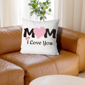 A square white throw pillow with a "MOM I Love You" design, where the "O" is replaced by a large pink heart, placed on a brown leather sofa.