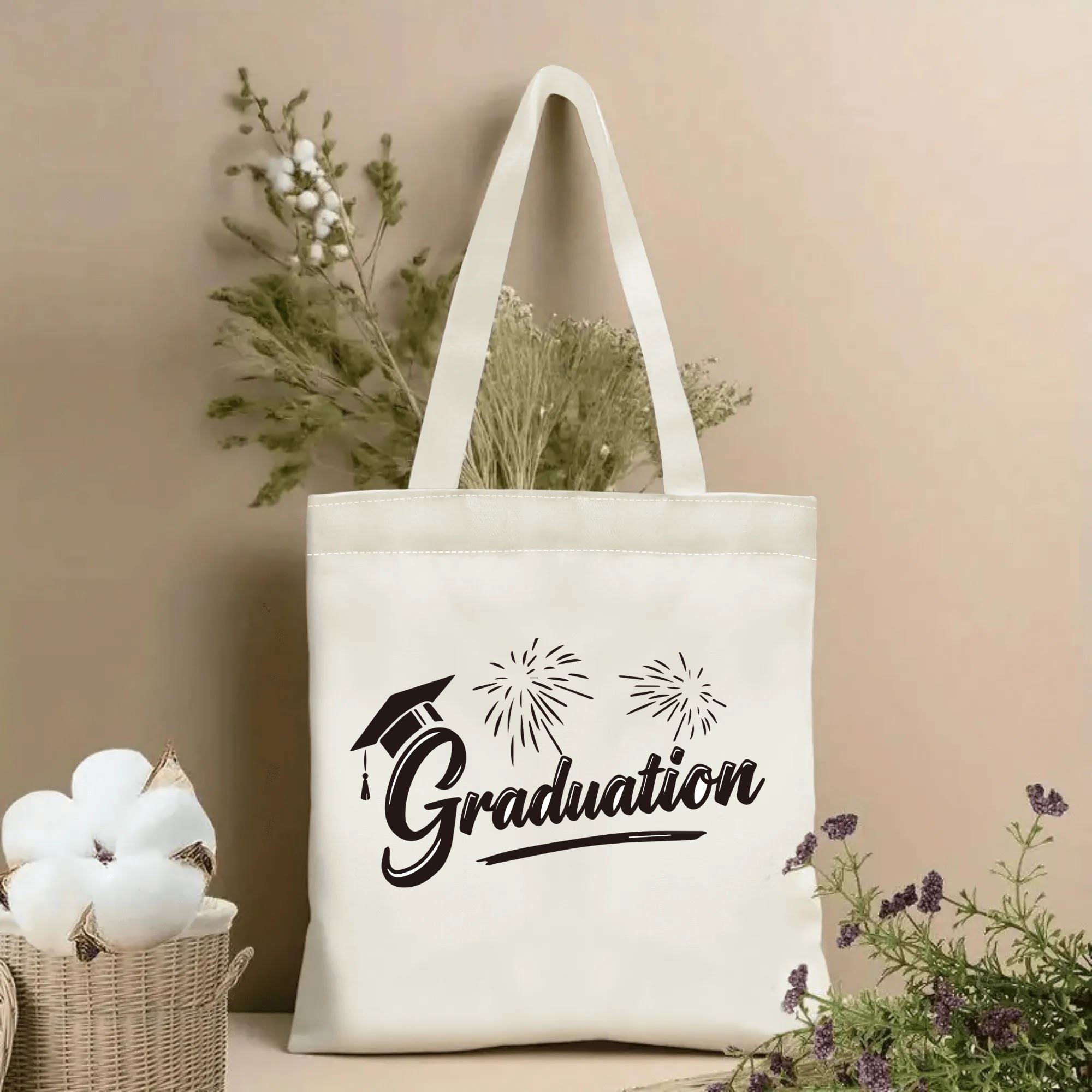 Natural canvas graduation‑themed tote bag featuring cursive "Graduation" lettering, graduation cap and fireworks print, styled with dried flowers for graduation gift display