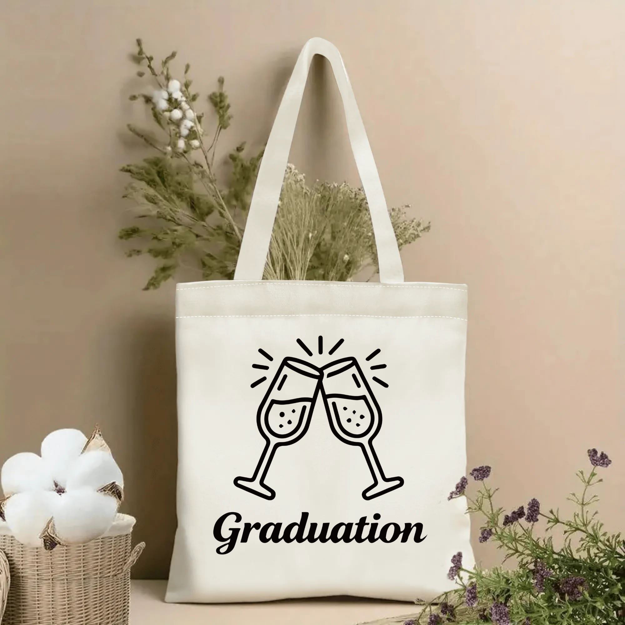 Natural canvas graduation‑themed tote bag featuring cursive "Graduation" lettering and clinking champagne glasses print, styled with dried flowers for graduation gift display