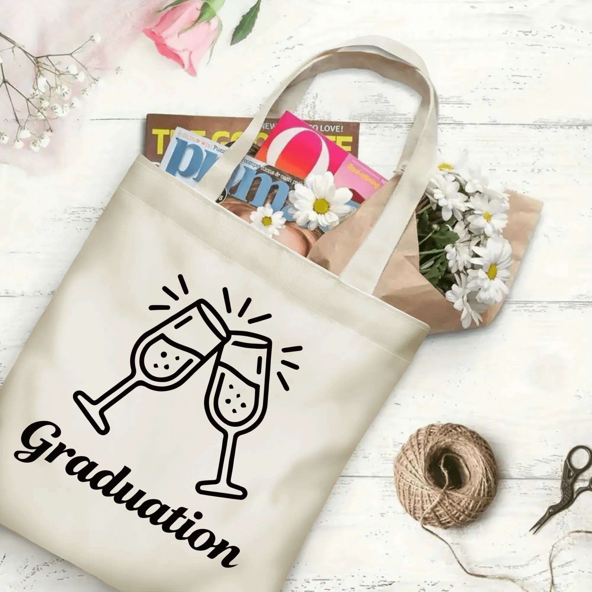 Flat‑lay shot of natural canvas graduation tote bag with champagne glasses and "Graduation" cursive print, decorated with daisies and books, ideal graduation gift