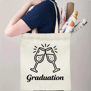 Person carrying natural canvas graduation tote bag with clinking champagne glasses and "Graduation" cursive print, casual celebration‑style reusable gift bag