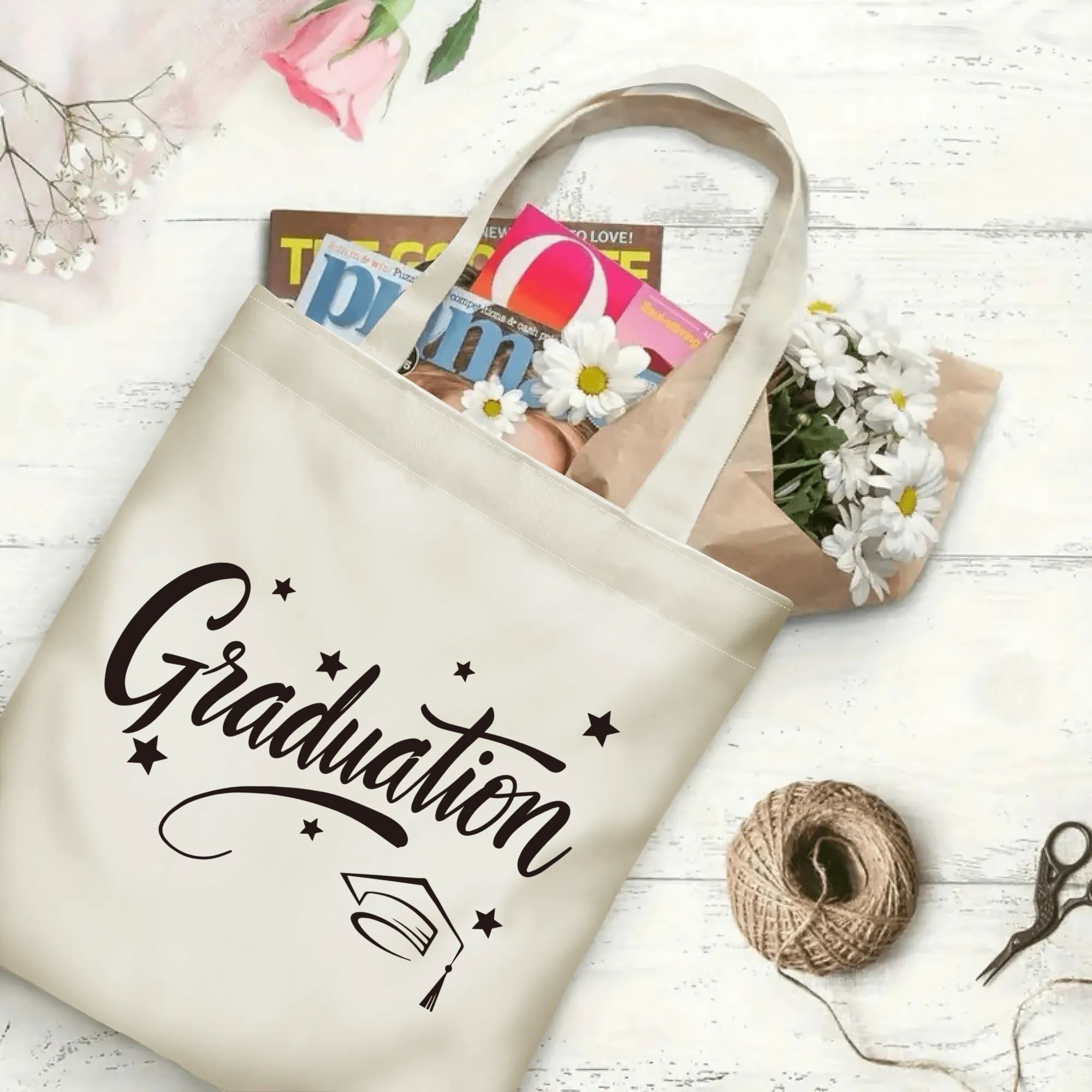 Flat‑lay shot of natural canvas graduation tote bag with flowing cursive "Graduation", graduation cap and star accents, decorated with daisies and books, ideal graduation gift