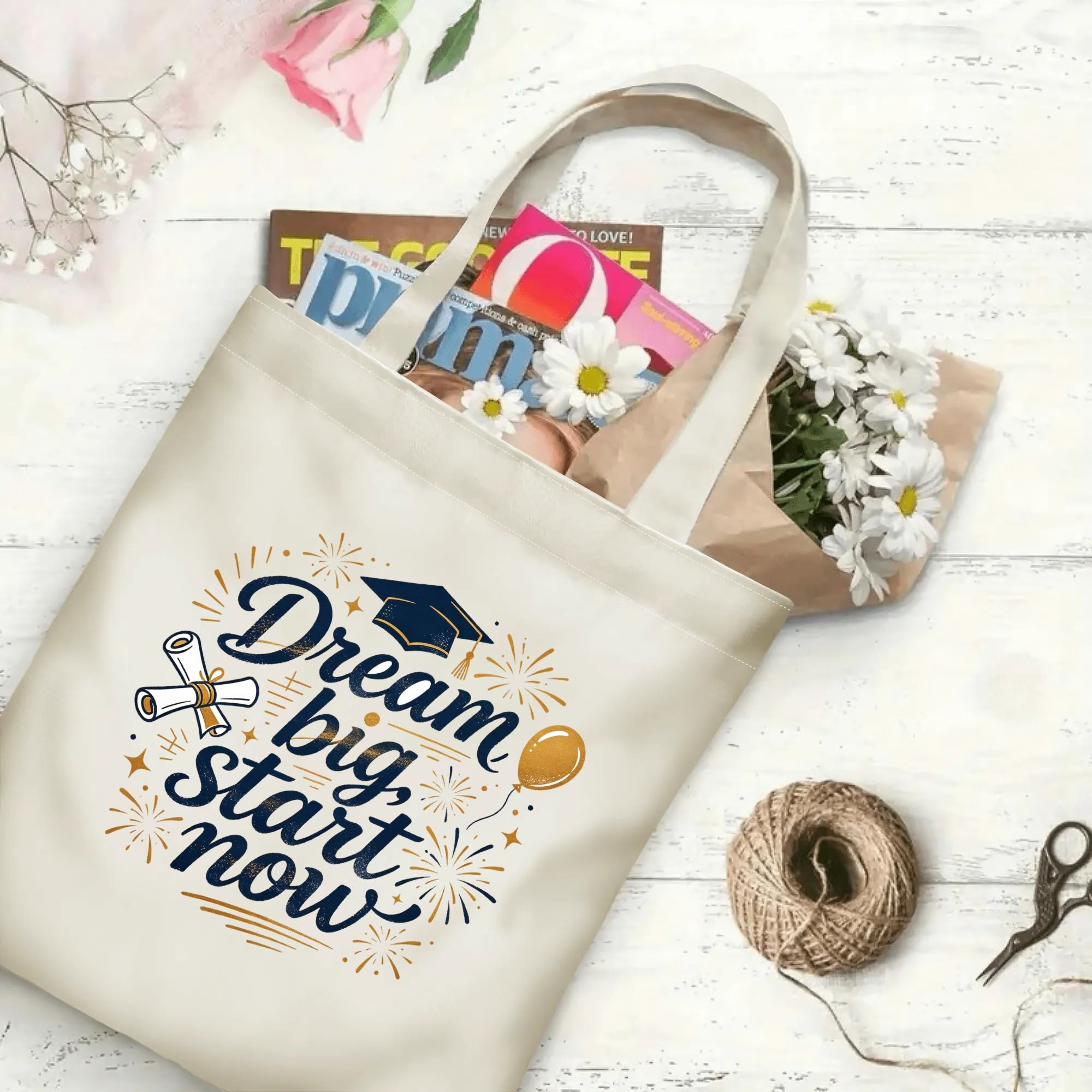 Flat‑lay shot of natural canvas graduation tote bag with inspirational "Dream big, start now" lettering, graduation cap, diploma, balloon and fireworks accents, decorated with daisies and books, ideal graduation gift