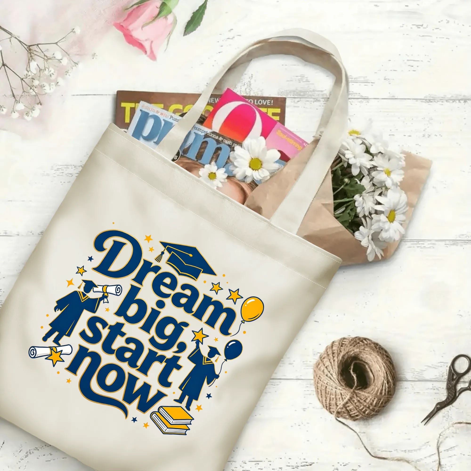 Flat‑lay shot of natural canvas graduation tote bag with inspirational "Dream big, start now" lettering, graduate figures, graduation cap, books and star details, decorated with daisies and books, ideal graduation gift