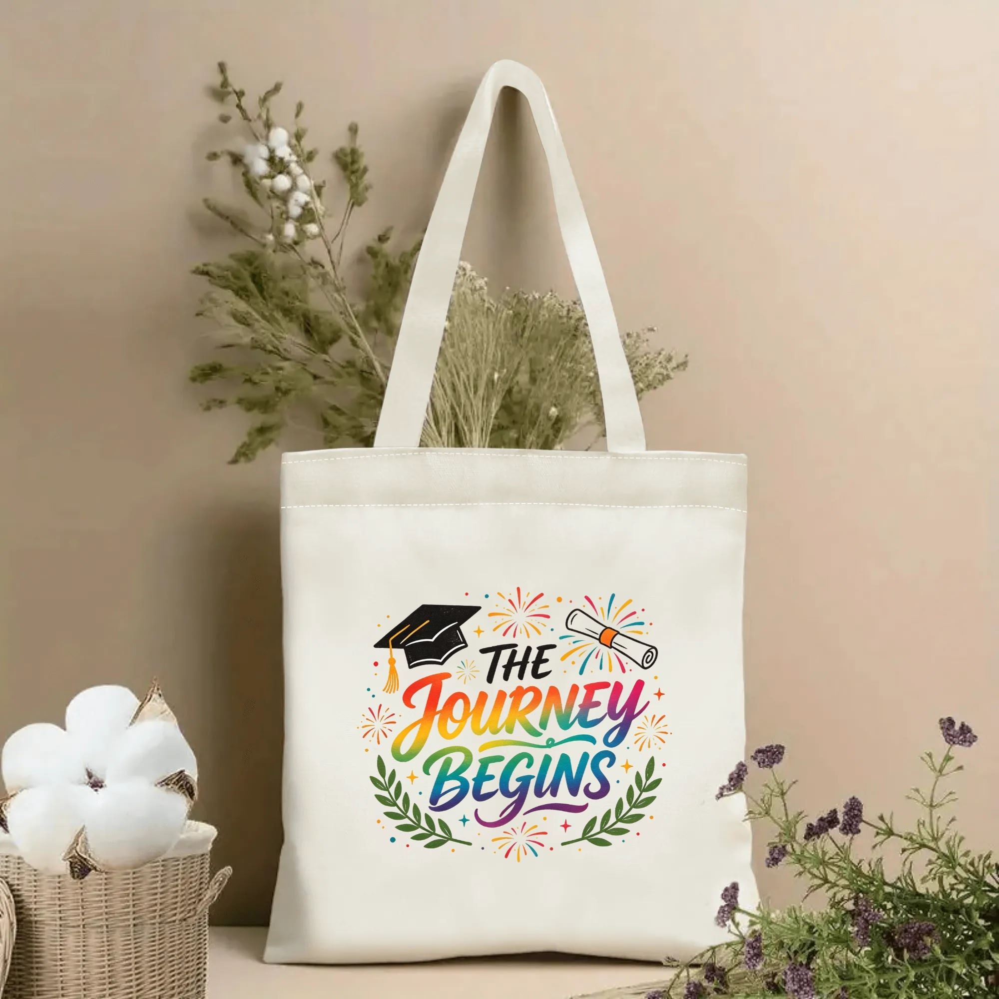 Natural canvas graduation‑themed tote bag featuring "The Journey Begins" rainbow‑gradient cursive quote, decorated with graduation cap, diploma, fireworks and laurel wreath, styled with dried flowers for graduation gift display
