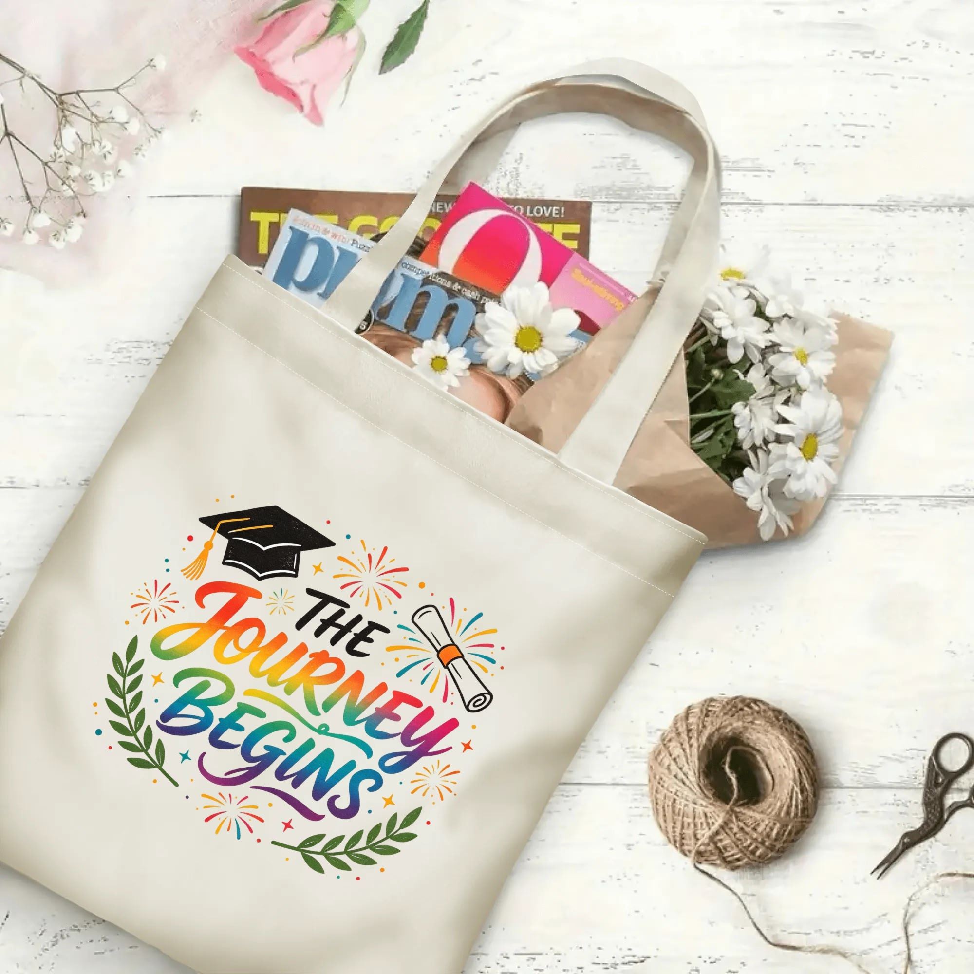 Flat‑lay shot of natural canvas graduation tote bag with vibrant rainbow‑gradient "The Journey Begins" print, graduation cap, diploma and celebratory details, decorated with daisies and books, ideal graduation gift