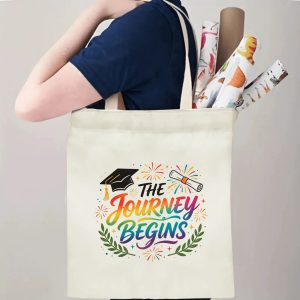 Person carrying natural canvas graduation tote bag with inspirational "The Journey Begins" rainbow‑colored print, graduation cap and fireworks graphics, casual milestone‑celebrating reusable gift bag