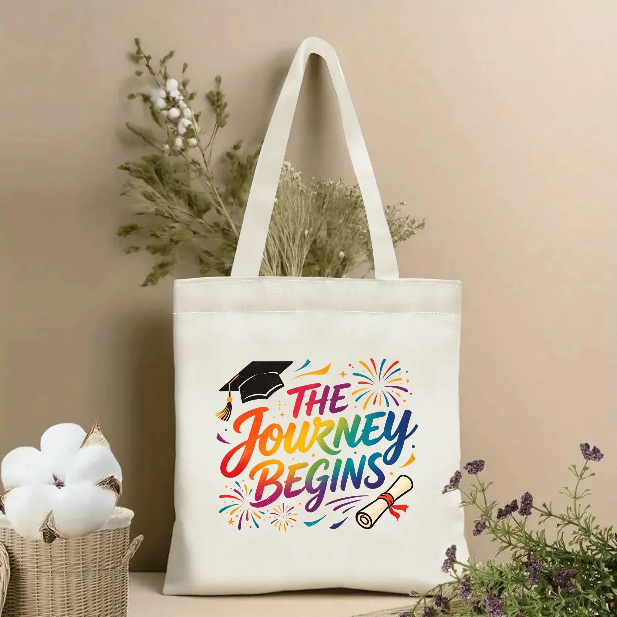 Natural canvas graduation‑themed tote bag featuring "The Journey Begins" rainbow‑gradient cursive lettering, decorated with graduation cap, diploma and vibrant fireworks accents, styled with dried flowers for graduation gift display