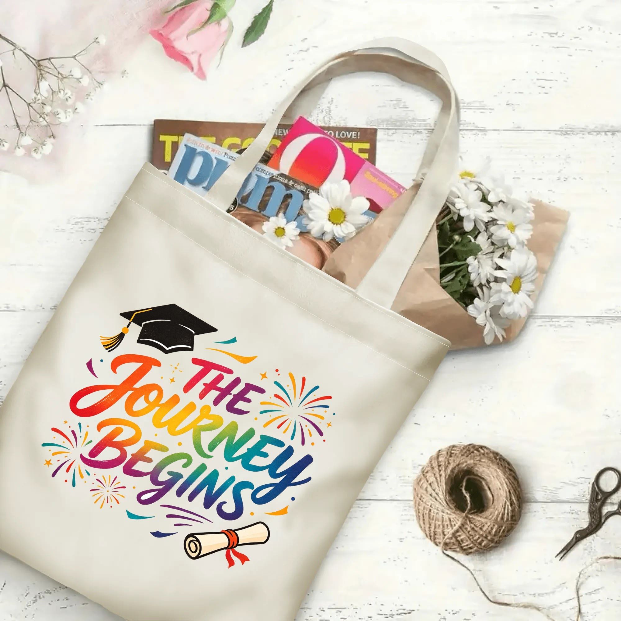 Flat‑lay shot of natural canvas graduation tote bag with colorful rainbow‑gradient "The Journey Begins" inspirational print, graduation cap, diploma and fireworks details, decorated with daisies and books, ideal graduation gift