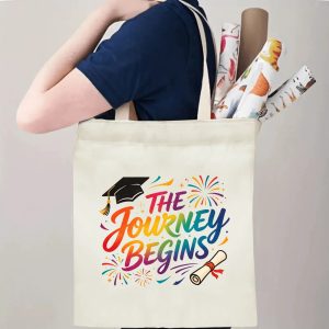 Person carrying natural canvas graduation tote bag with celebratory rainbow‑colored "The Journey Begins" print, graduation cap and diploma graphics, casual milestone‑celebrating reusable gift bag