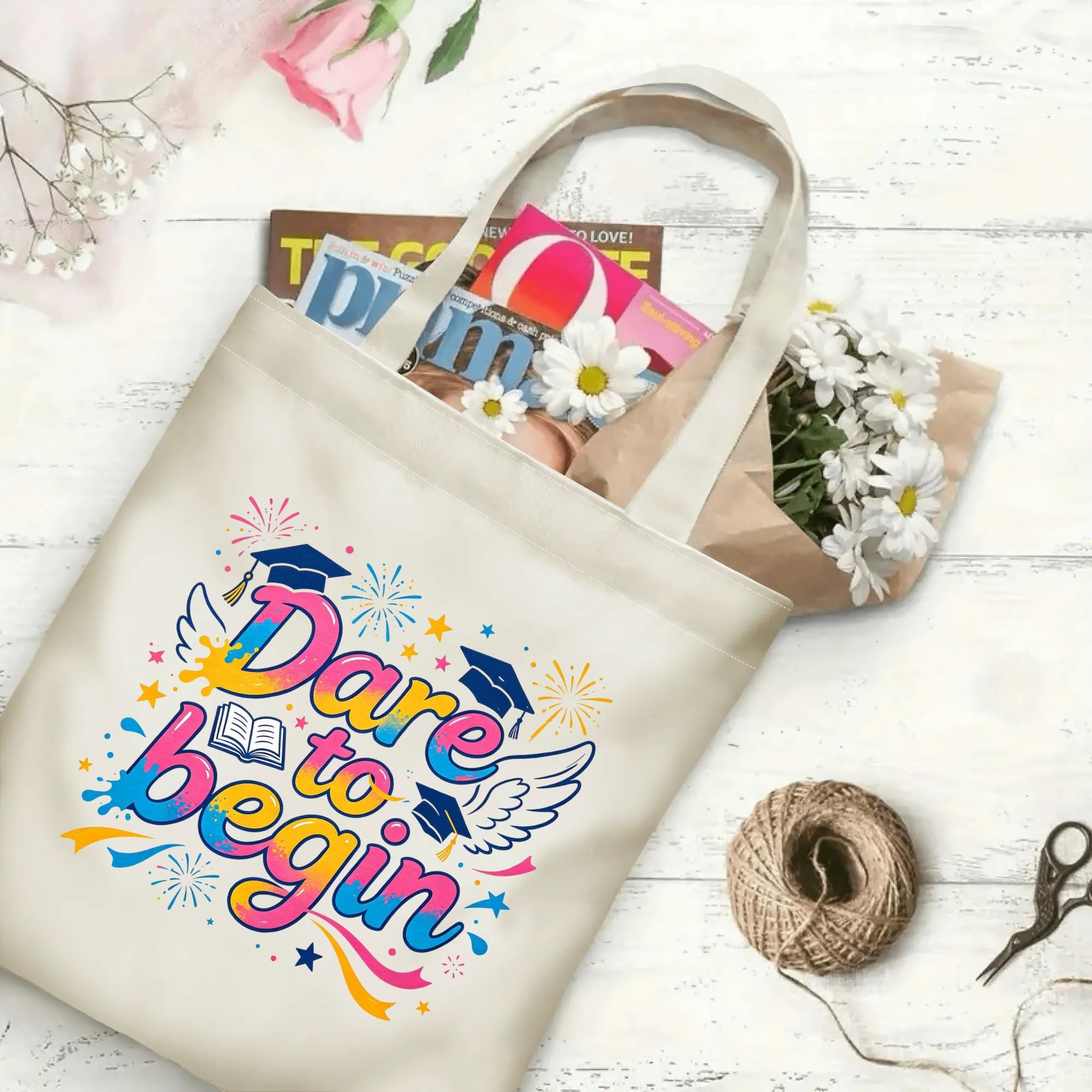 Flat‑lay shot of natural canvas graduation tote bag with bold colorful "Dare to begin" inspirational print, graduation caps, wings, book and celebratory details, decorated with daisies and books, ideal graduation gift