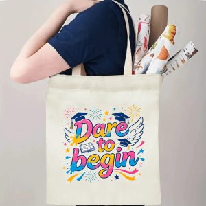 Person carrying natural canvas graduation tote bag with energetic "Dare to begin" rainbow‑colored print, graduation cap and wing graphics, casual milestone‑celebrating reusable gift bag