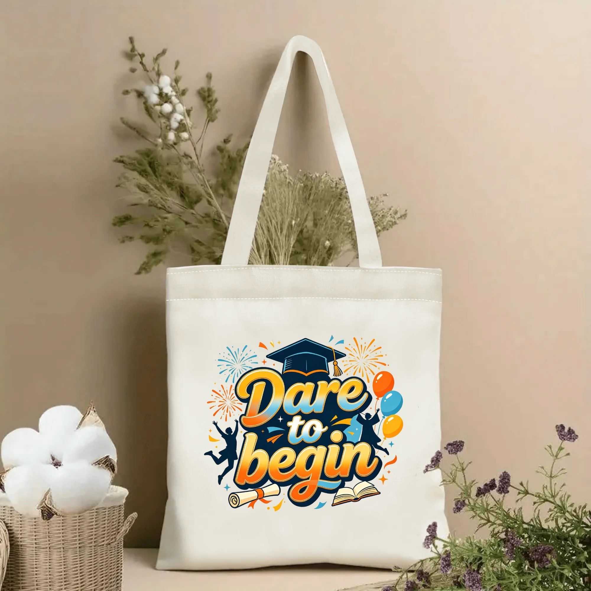 Natural canvas graduation‑themed tote bag featuring "Dare to begin" gradient cursive lettering, decorated with graduation cap, diploma, open book, cheering graduate silhouettes, balloons and fireworks, styled with dried flowers for graduation gift display