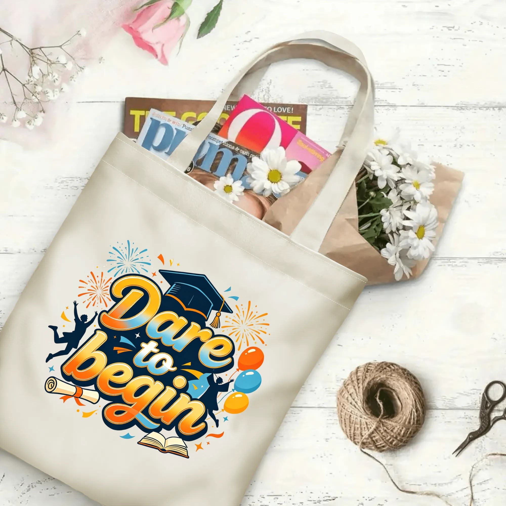 Flat‑lay shot of natural canvas graduation tote bag with bold gradient "Dare to begin" motivational print, graduation cap, graduates, balloons and celebratory details, decorated with daisies and books, ideal graduation gift