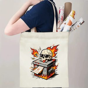 Person carrying natural canvas 2026 mid‑term election gag gift tote bag with fiery skull ballot box tattoo‑style design and edgy punk‑rock aesthetic, reusable political accessory for election rallies, voting day and everyday errands