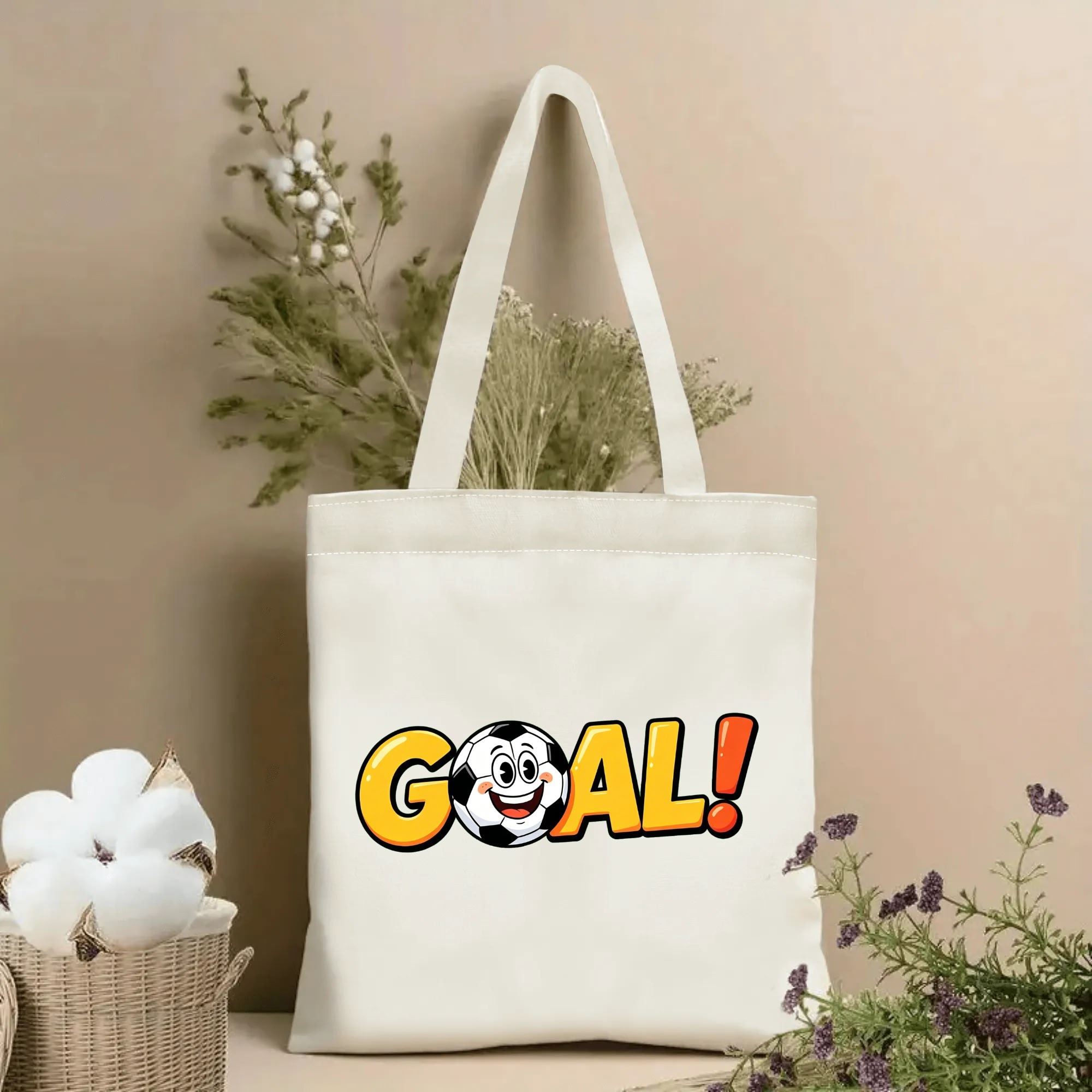 Natural canvas soccer‑themed tote bag featuring playful cartoon "GOAL!" lettering with a smiling soccer ball character integrated into the design, styled with dried flowers for sports‑fan gift display