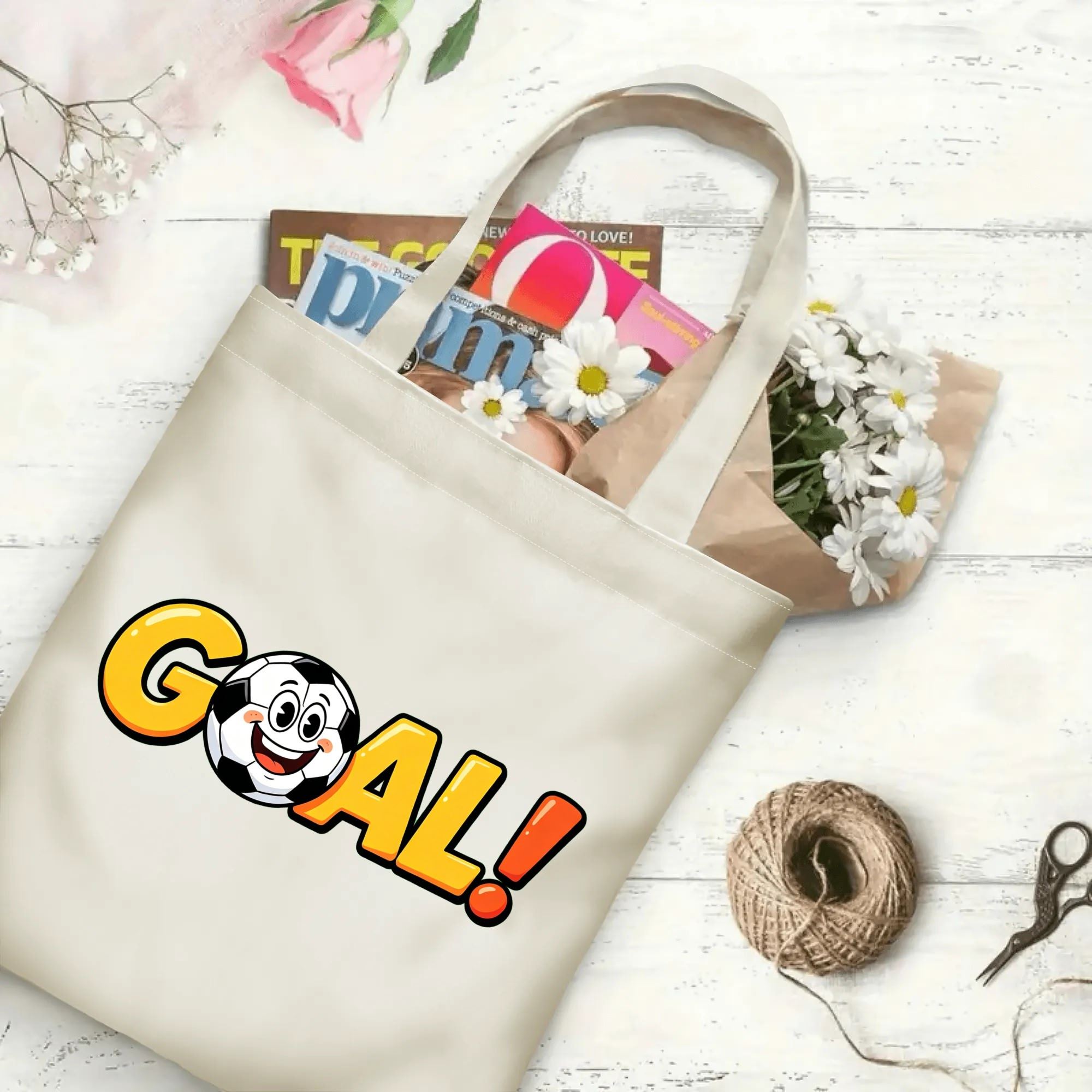 Flat‑lay shot of natural canvas sports novelty tote bag with fun bold "GOAL!" typography and cute happy soccer‑ball graphic, decorated with daisies, ideal casual daily‑wear bag for football fans