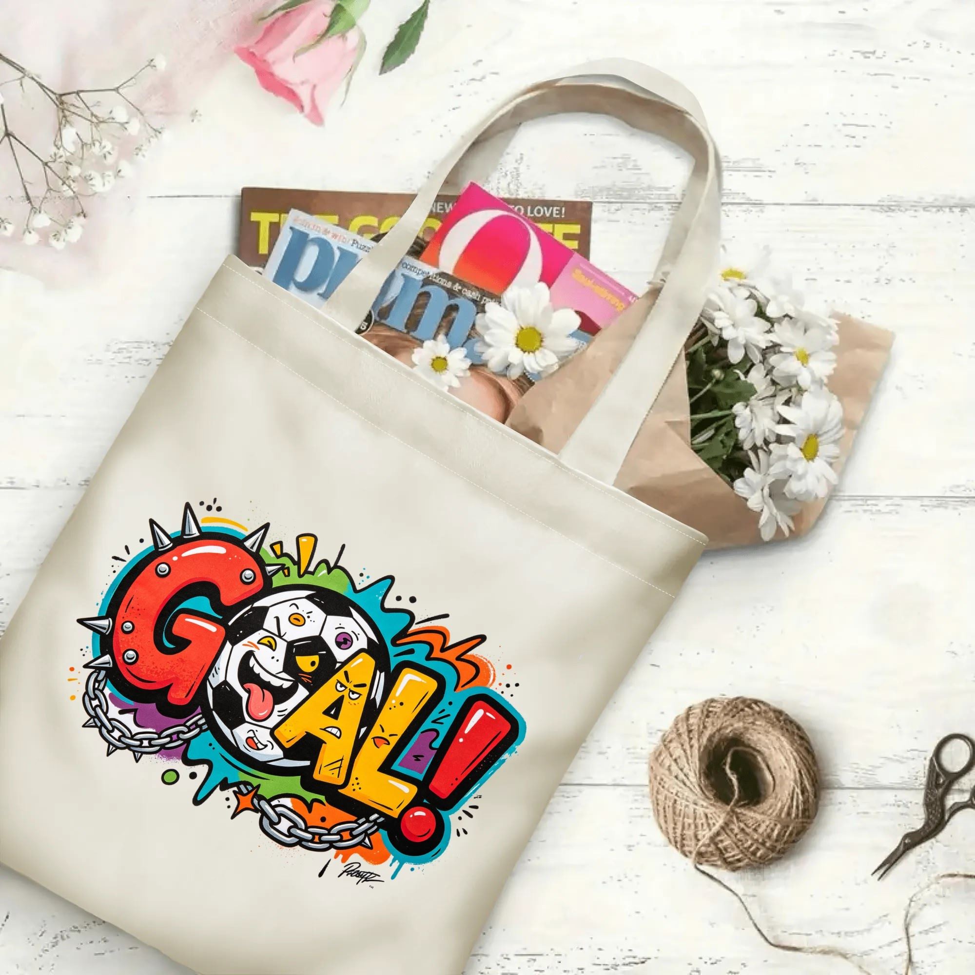Flat‑lay shot of natural canvas street‑art sports novelty tote bag with bold colorful punk‑style "GOAL!" typography, grunge splatter details and cheeky soccer‑ball mascot, decorated with daisies, ideal casual daily‑wear bag for football fans
