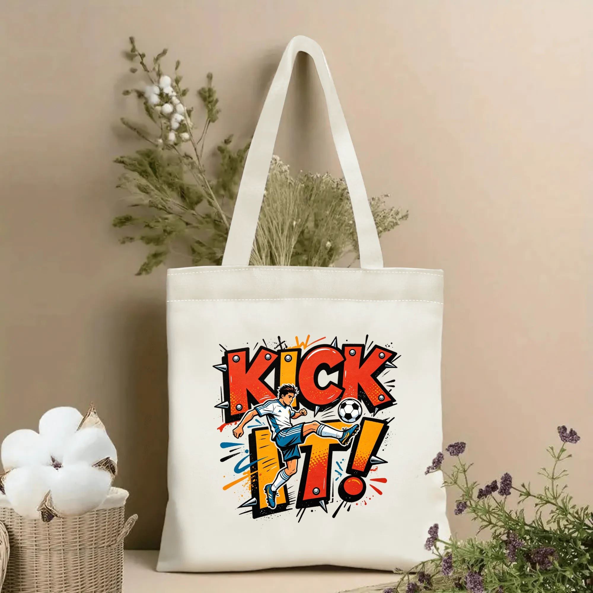 Natural canvas soccer‑themed tote bag featuring an edgy punk‑graffiti style "KICK IT!" spiked lettering graphic, a kicking soccer player cartoon and colorful splatter‑paint accents, styled with dried flowers for sports‑fan gift display