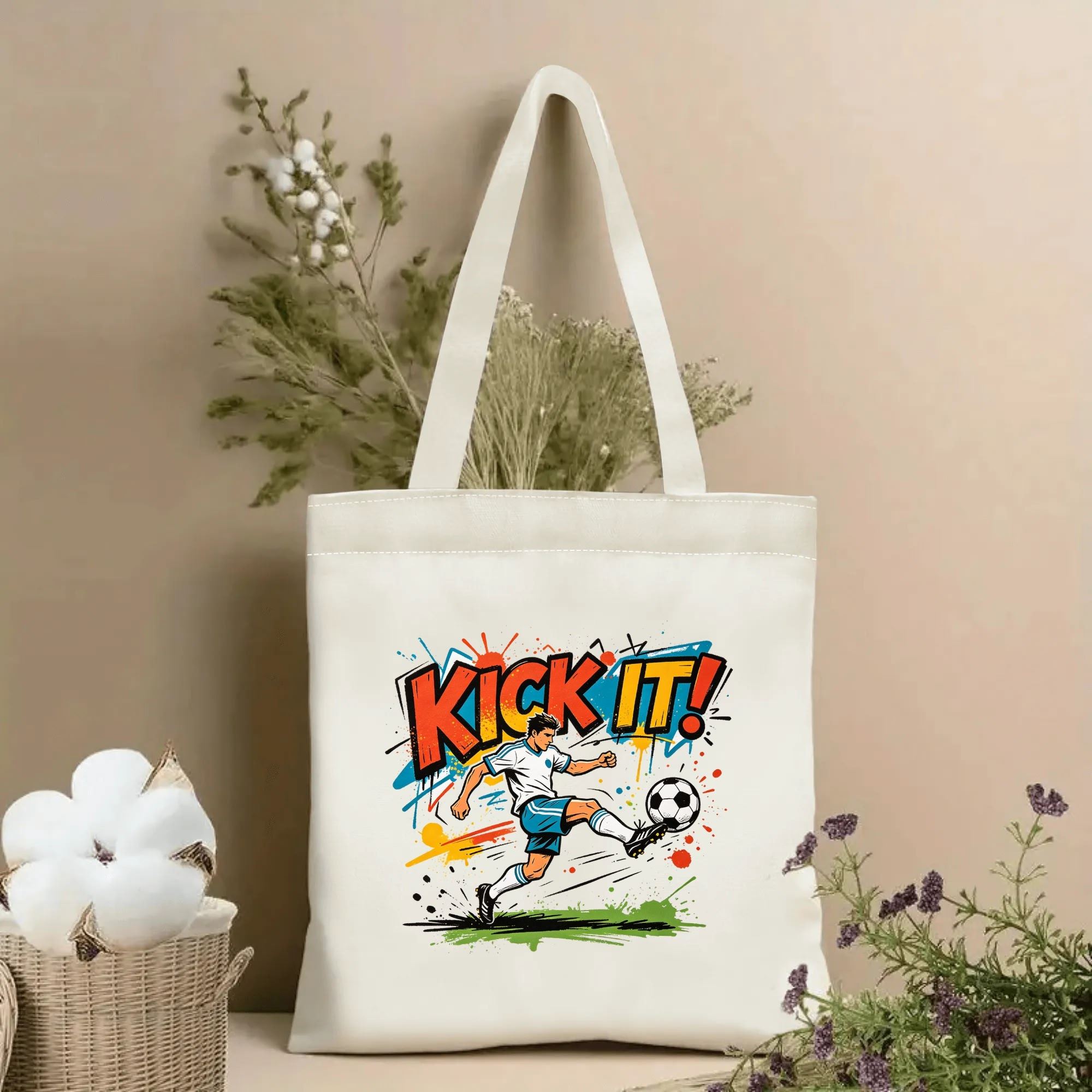 Natural canvas soccer‑themed tote bag featuring vibrant street‑graffiti style "KICK IT!" bold lettering, dynamic kicking soccer player graphic and colorful paint‑splatter splash accents, styled with dried flowers for sports‑fan gift display