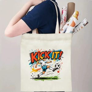 Person carrying natural canvas trendy soccer fan‑gift tote bag with bright graffiti‑pop "KICK IT!" design and action‑packed soccer‑player mascot, reusable sports accessory for match days, fan events or everyday errands