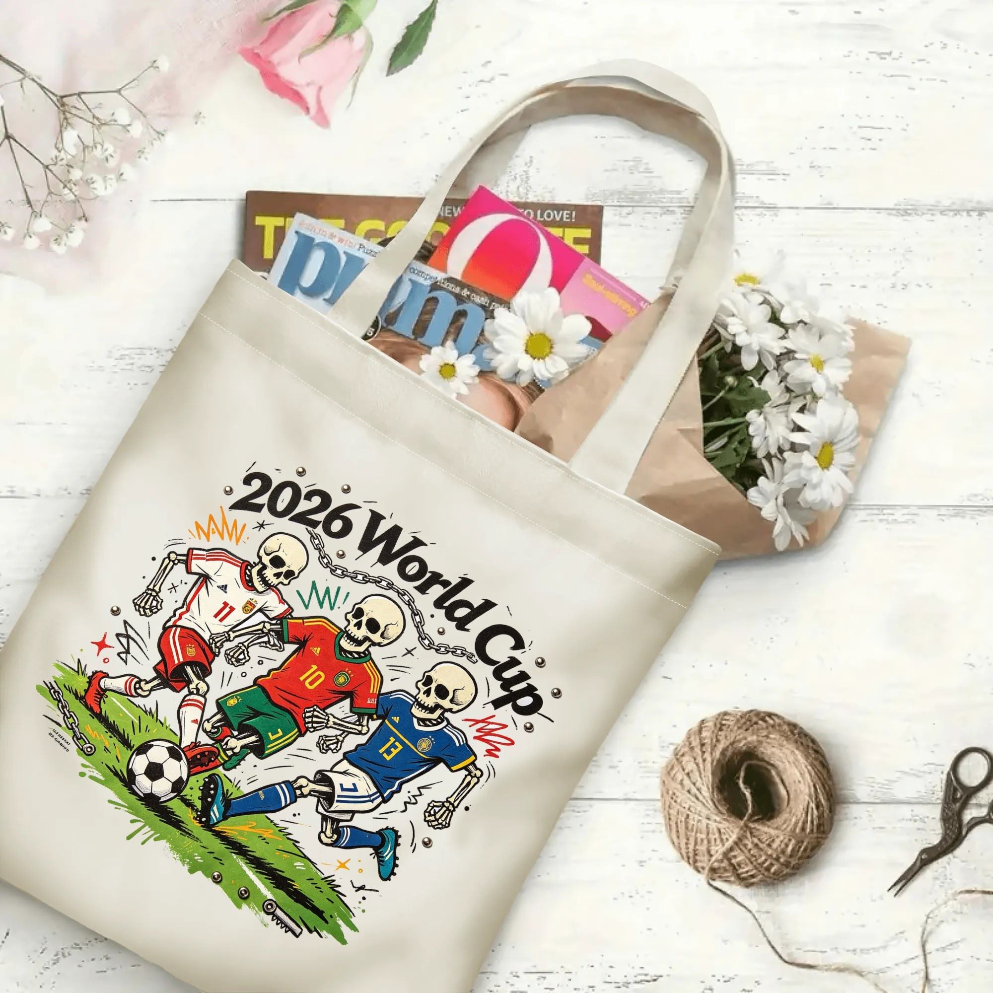 Flat‑lay shot of natural canvas novelty football tote bag with bold street‑art skeleton soccer graphic, three skull players in red‑white, red‑green and blue kits and chaotic punk‑style field elements, decorated with daisies, ideal casual daily‑wear bag for World Cup fans