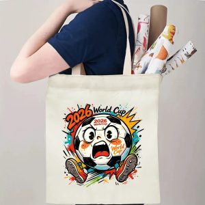 Person carrying natural canvas 2026 World Cup fan‑gift tote bag with lively expressive jumping soccer‑ball mascot design, reusable sports accessory for tournament viewing, fan events or everyday errands