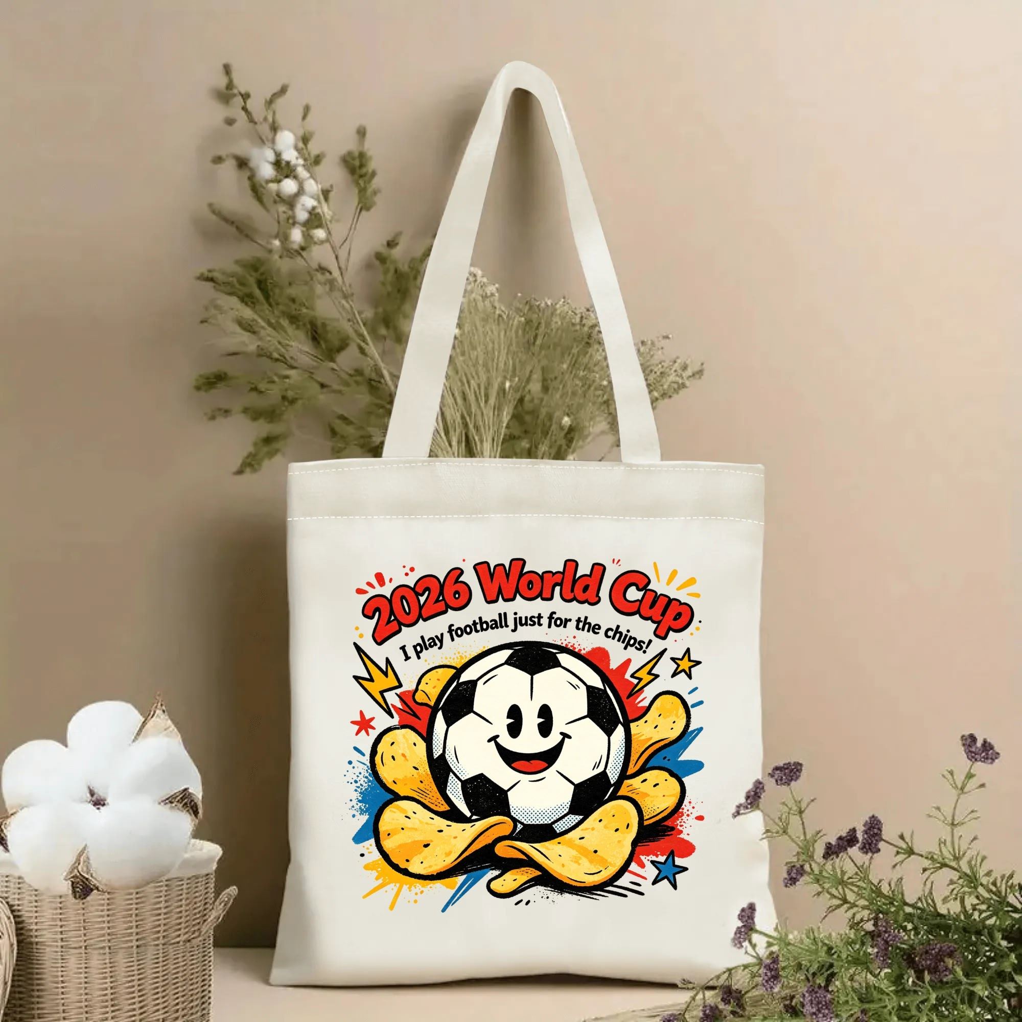 Natural canvas 2026 World Cup‑themed tote bag featuring a fun cartoon‑pop graphic of a smiling anthropomorphic soccer‑ball mascot surrounded by crispy potato chips, playful lightning‑star accents, colorful paint‑splatter details and humorous quote “I play football just for the chips!”, styled with dried flowers for sports‑fan gift display