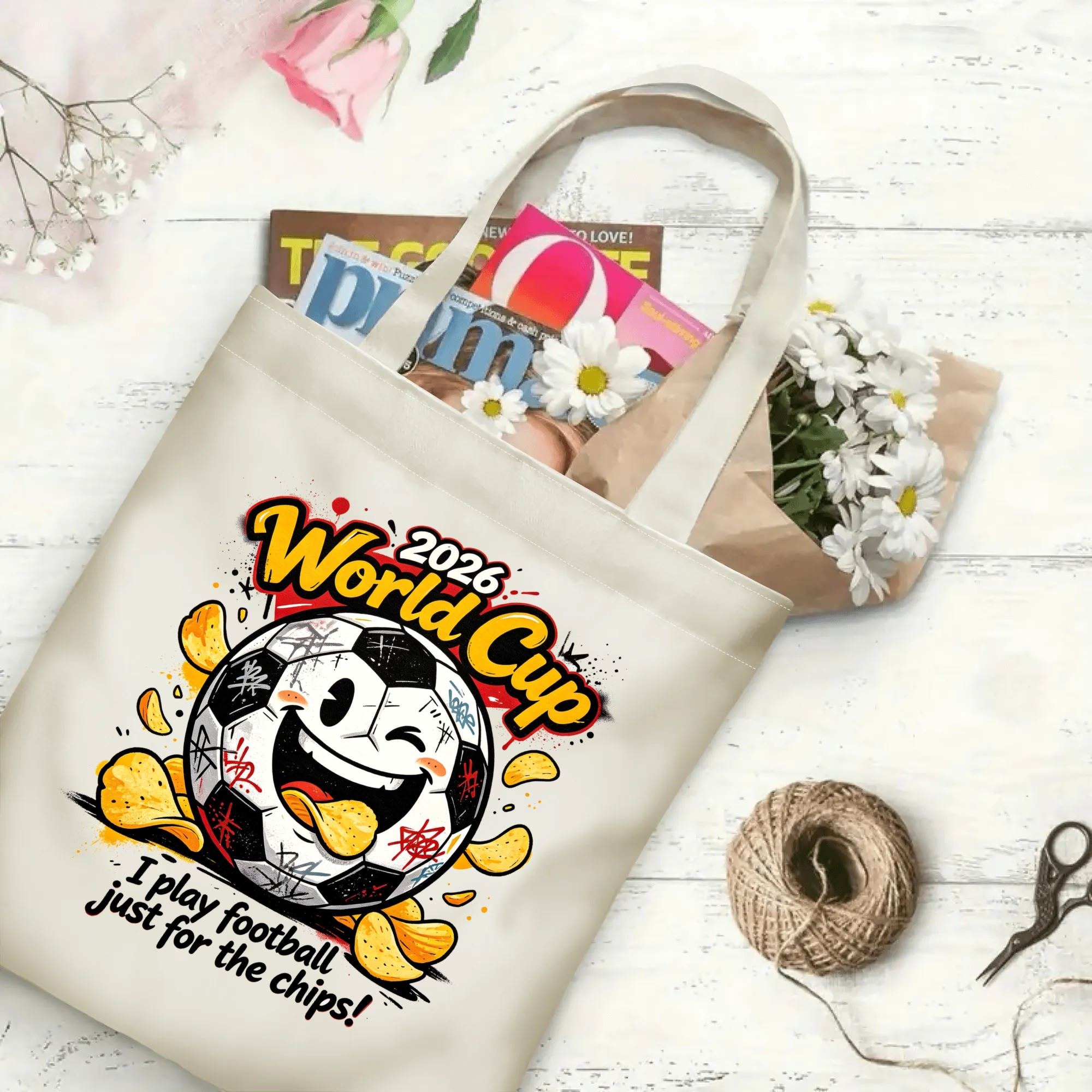 Flat‑lay shot of natural canvas novelty football tote bag with edgy grunge soccer‑and‑chips cartoon graphic, cheeky winking soccer‑ball character and funny snack‑humor quote, decorated with daisies, ideal casual daily‑wear bag for World Cup fans