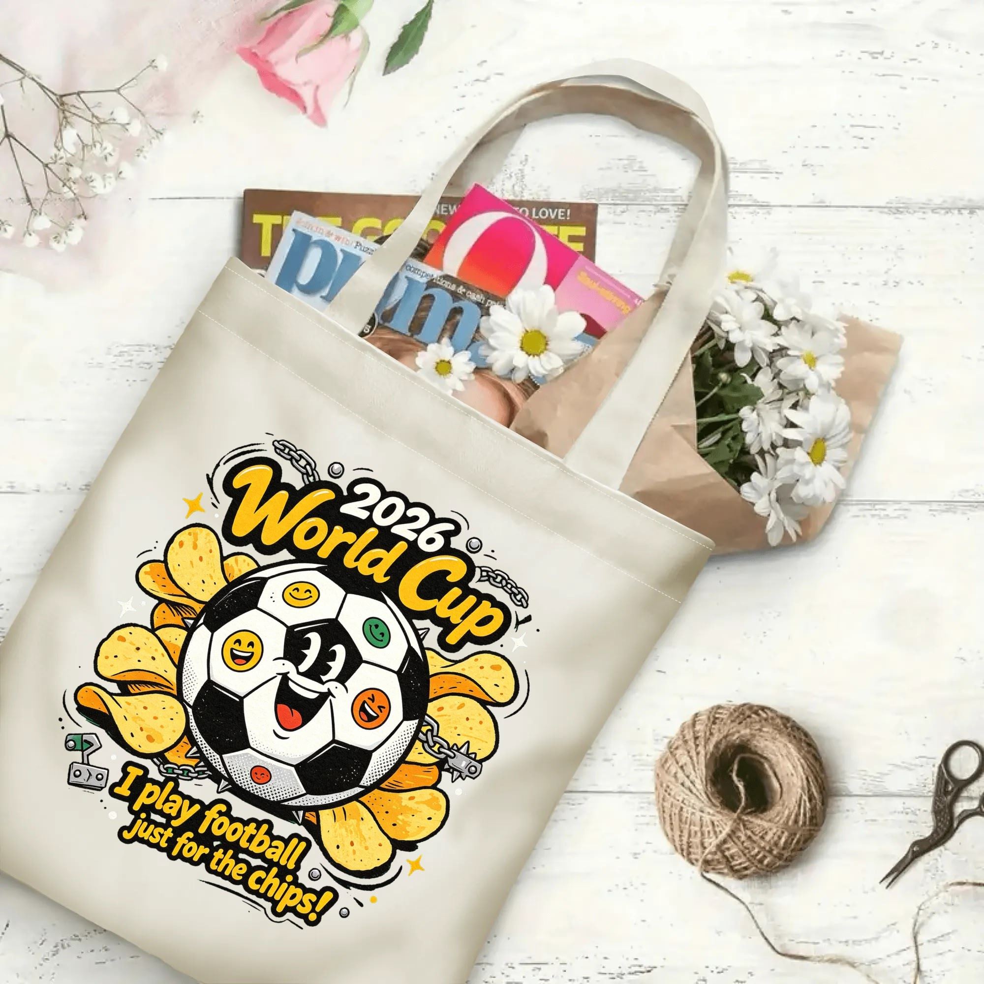 Flat‑lay shot of natural canvas novelty football tote bag with fun grunge punk soccer‑and‑chips cartoon graphic, cheerful emoji‑faced soccer‑ball character and witty snack‑humor quote, decorated with daisies, ideal casual daily‑wear bag for World Cup fans