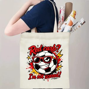 Person carrying natural canvas soccer fan‑gift tote bag with rebellious cool street‑cartoon soccer‑ball mascot design, reusable sports accessory for match days, fan events or everyday errands