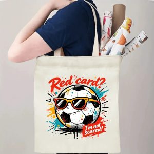 Person carrying natural canvas soccer fan‑gift tote bag with cool laid‑back street‑cartoon soccer‑ball mascot design, reusable sports accessory for match days, fan events or everyday errands