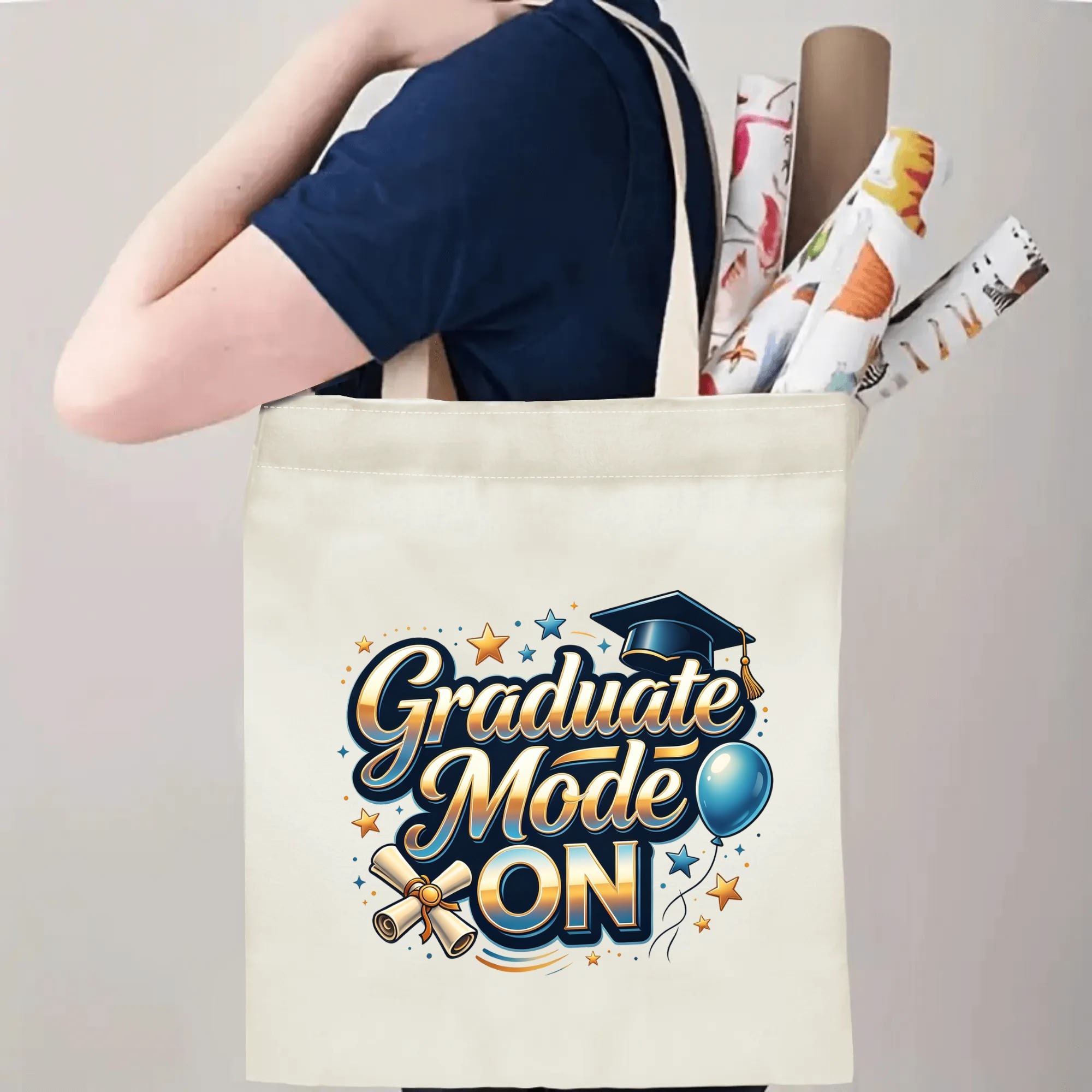 Person carrying a "Graduate Mode ON" canvas tote bag over the shoulder, holding rolled posters and graduation essentials
