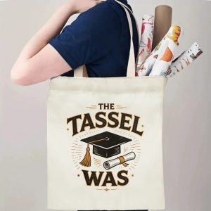 Person carrying a “THE TASSEL WAS” graduation tote bag over the shoulder, with rolled posters and graduation essentials.