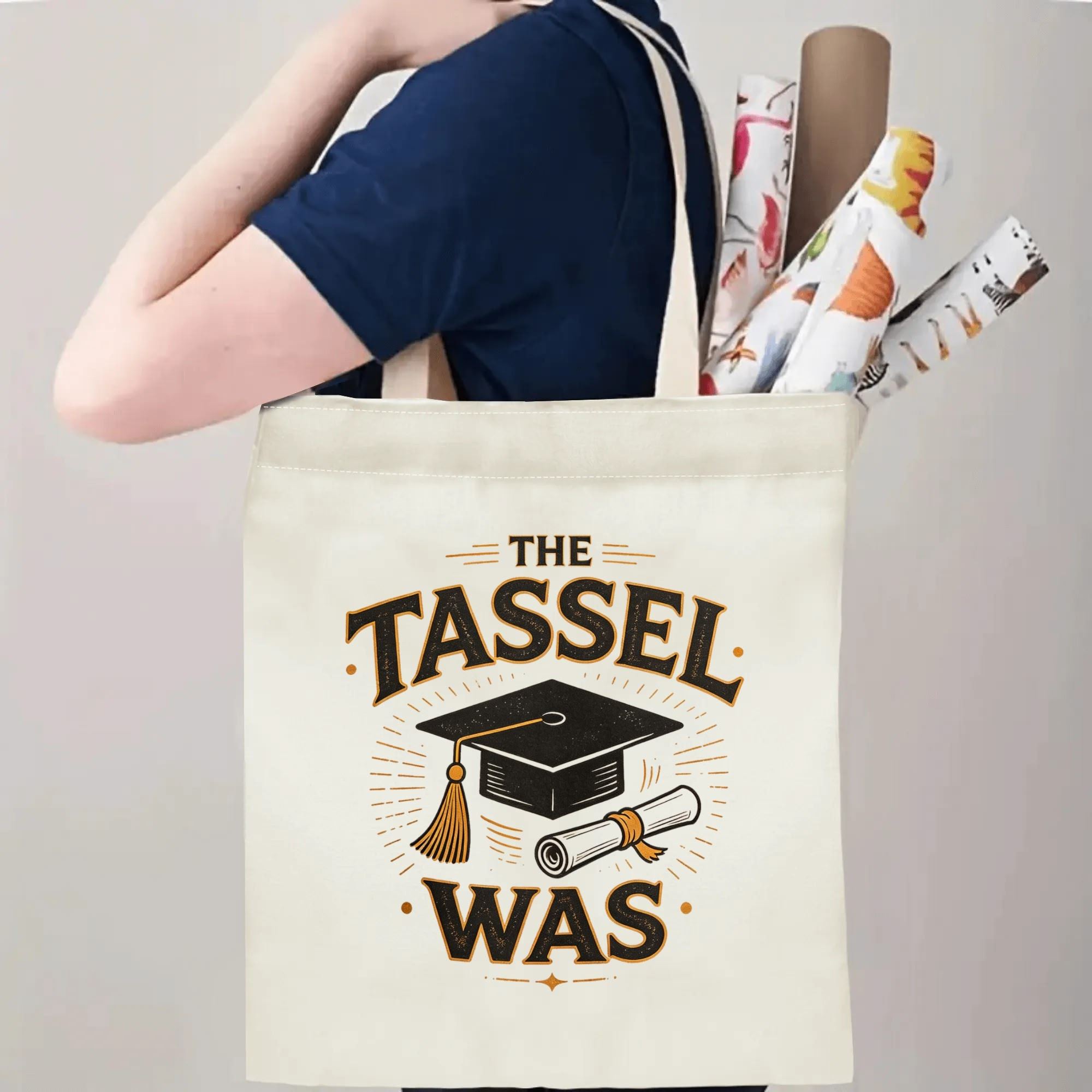 Person carrying a “THE TASSEL WAS” graduation tote bag over the shoulder, with rolled posters and graduation essentials.