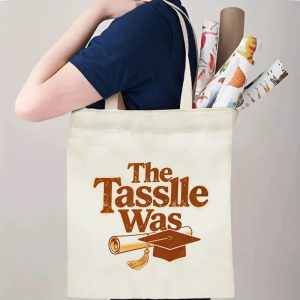 Person carrying a “The Tassle Was” graduation tote bag over the shoulder, with rolled posters and graduation essentials.